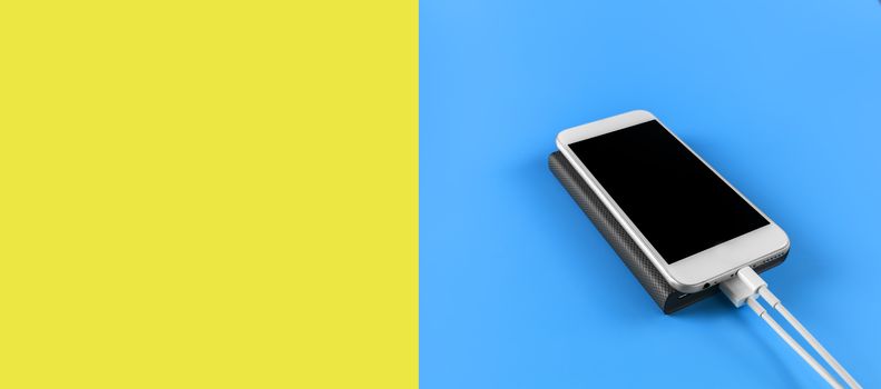 Top view smartphone charging with power bank on blue and empty yellow background with copy space. Smartphone charging with power bank by white USB cable.