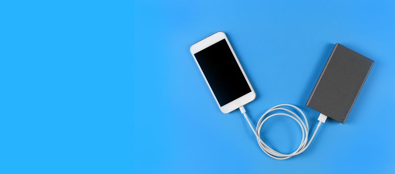 Top view smartphone charging with power bank on blue background with copy space. Smartphone charging with power bank by white USB cable.