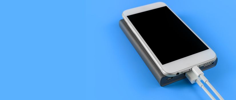 Close up smartphone charging with power bank on blue background with copy space. Smartphone charging with power bank by white USB cable.