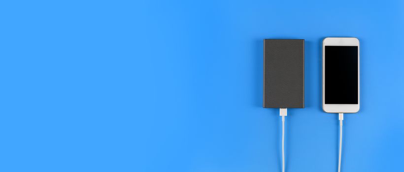 Top view smartphone charging with power bank on blue background with copy space. Smartphone charging with power bank by white USB cable.