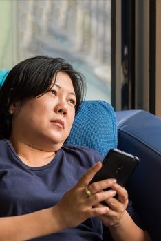 Asian women 40s white skin and plump body in blue dress holding smartphone have a doubt and think gesture on a blue sofa near window