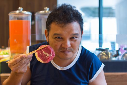 Asia man plump body holding a beef with chopsticks, defocus blurred on BBQ. yakiniku grill buffet restaurant background.