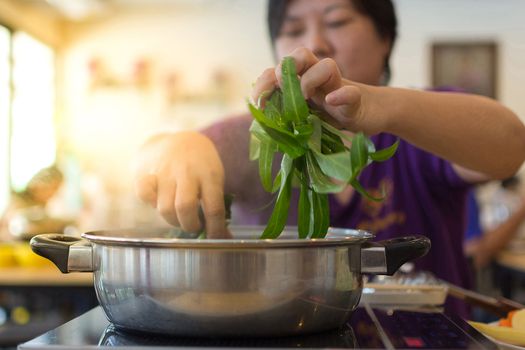 Asia woman plump body cooking food by put a vegetable (morning glory) in to a hot pot for make a sukiyaki or shabu , process in soft orange sun light style