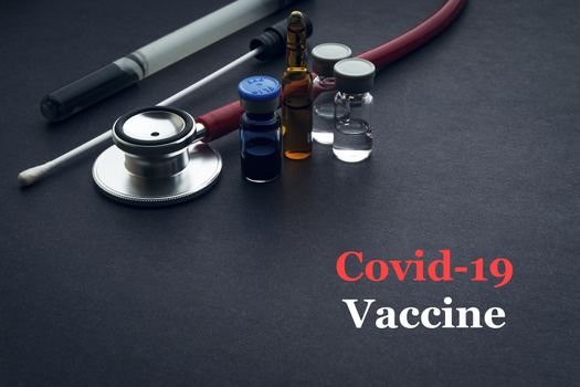 COVID-19 or CORONAVIRUS VACCINE text with stethoscope, medical swab and vial on black background. Covid-19 or Coronavirus concept. 