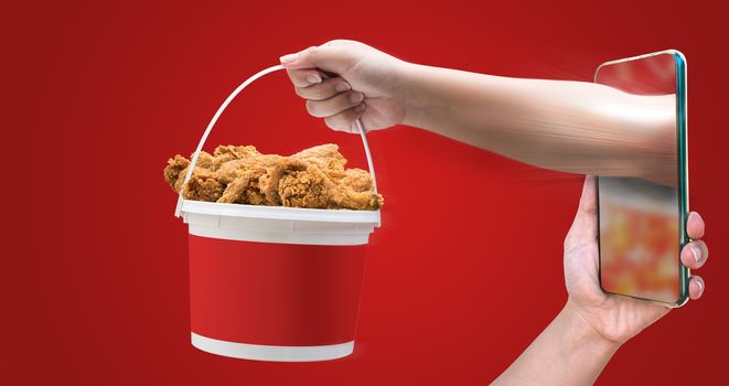 Fantasy pictures showing food ordering via mobile phones And the hand holding a bucket of crispy fried chicken through the mobile phone screen on the red background