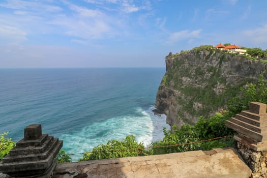 View of Uluwatu cliff with pavilion and blue sea in Bali, Indonesia. Beautiful scenery of Pura Luhur Uluwatu Temple with colorful flowers foreground during day. Scenic coastal landscape of high cliff.