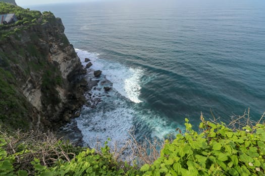 View of Uluwatu cliff with pavilion and blue sea in Bali, Indonesia. Beautiful scenery of Pura Luhur Uluwatu Temple with colorful flowers foreground during day. Scenic coastal landscape of high cliff.