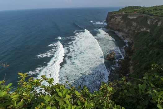 View of Uluwatu cliff with pavilion and blue sea in Bali, Indonesia. Beautiful scenery of Pura Luhur Uluwatu Temple with colorful flowers foreground during day. Scenic coastal landscape of high cliff.