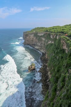 View of Uluwatu cliff with pavilion and blue sea in Bali, Indonesia. Beautiful scenery of Pura Luhur Uluwatu Temple with colorful flowers foreground during day. Scenic coastal landscape of high cliff.