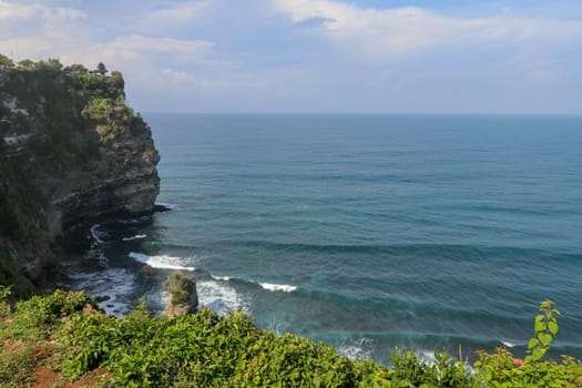 Overview panorama ocean shore, cliff. Overwhelmed scene the flower and green-capped vertical cliff over the Indian ocean. South of Bali island, Pura Luhur Uluwatu Temple. Indonesia.