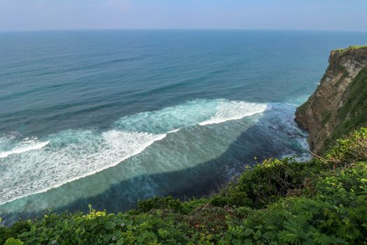 Overview panorama ocean shore, cliff. Overwhelmed scene the flower and green-capped vertical cliff over the Indian ocean. South of Bali island, Pura Luhur Uluwatu Temple. Indonesia.