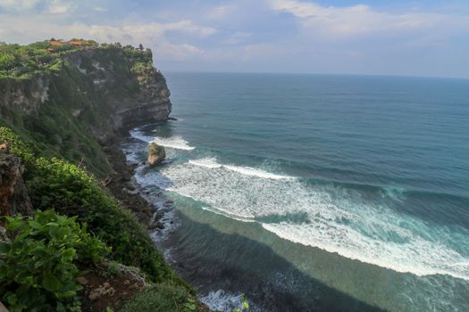 Overview panorama ocean shore, cliff. Overwhelmed scene the flower and green-capped vertical cliff over the Indian ocean. South of Bali island, Pura Luhur Uluwatu Temple. Indonesia.