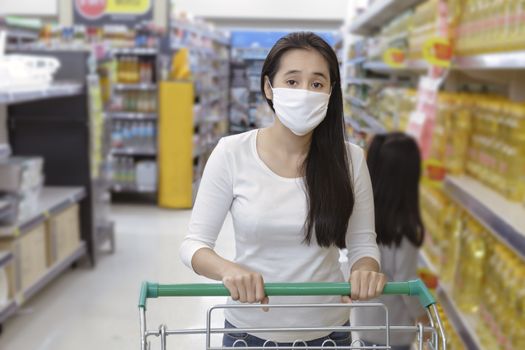 Asian woman wear face mask push shopping cart in suppermarket departmentstore. Girl choosing, looking grocery to buy  something. During coronavirus crisis or covid19 outbreak. Women wearing protective face mask hold shopping cart, New normal after covid epidemic
