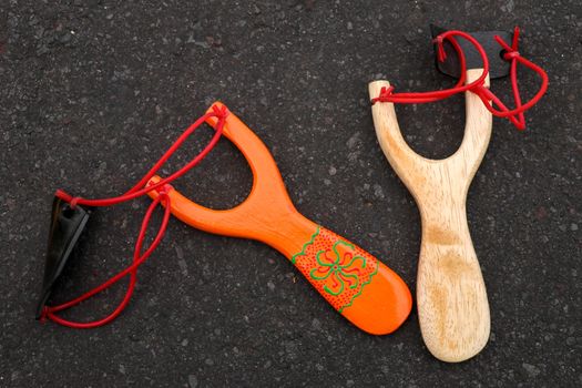 Couple of simple wooden slingshot isolated. Orange and natural wooden handle with two red rubber bands attached to a black leather strip used to put things to be shot on black background. Primitive but effective weapon.