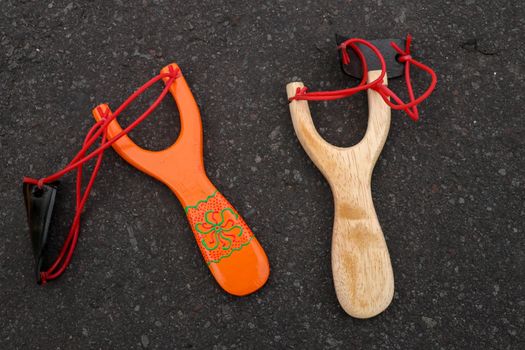 Couple of simple wooden slingshot isolated. Orange and natural wooden handle with two red rubber bands attached to a black leather strip used to put things to be shot on black background. Primitive but effective weapon.
