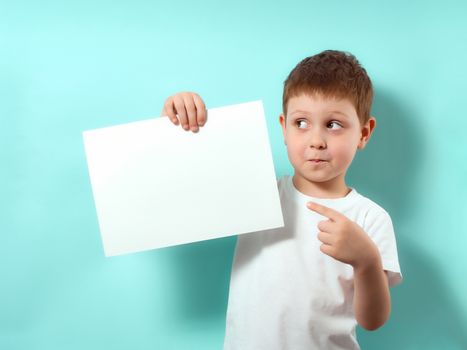 Four-year-old boy with a surprised face, looking away and shows finger on blank white sheet. Fun child on blue background with copy space for message, mock up