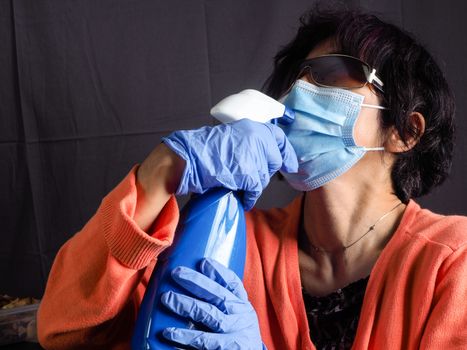 funny humorous adult asian 40s 50s woman wearing protection surgical face mask , blue latex gloves, and sun glasses pointing the head with blue sanitizer cleaning  bottle as a weapon as tired of confinement covid concept.