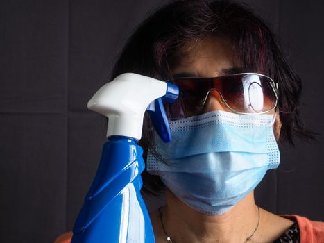 funny humorous adult asian 40s 50s woman wearing protection surgical face mask , blue latex gloves, and sun glasses pointing the head with blue sanitizer cleaning  bottle as a weapon as tired of confinement covid concept.