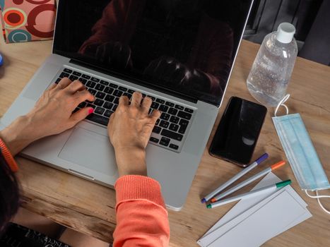 Woman hand s freelance working on a notebook laptop computer at home or home office with gel and face mask prevention items near connected to internet via hot spot of smartphone in fall, orange sweater and brown table, earth tones.
