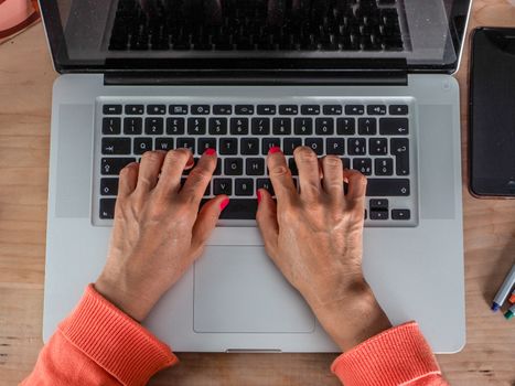 Woman hand s freelance working on a notebook laptop computer at home or home office with gel and face mask prevention items near connected to internet via hot spot of smartphone in fall, orange sweater and brown table, earth tones.