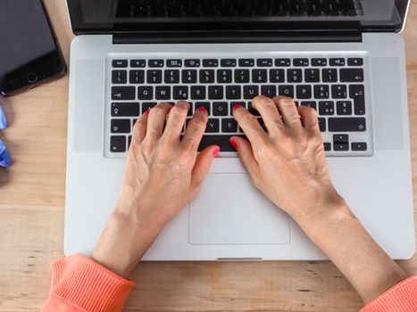 Woman hand s freelance working on a notebook laptop computer at home or home office with gel and face mask prevention items near connected to internet via hot spot of smartphone in fall, orange sweater and brown table, earth tones.