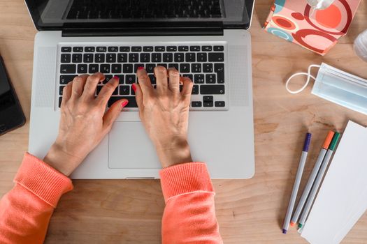 Woman hand s freelance working on a notebook laptop computer at home or home office with gel and face mask prevention items near connected to internet via hot spot of smartphone in fall, orange sweater and brown table, earth tones.