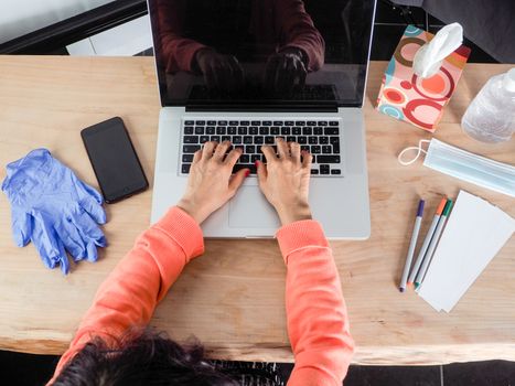 Woman hand s freelance working on a notebook laptop computer at home or home office with gel and face mask prevention items near connected to internet via hot spot of smartphone in fall, orange sweater and brown table, earth tones.