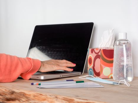 Woman hand s freelance working on a notebook laptop computer at home or home office with gel and face mask prevention items near connected to internet via hot spot of smartphone in fall, orange sweater and brown table, earth tones.