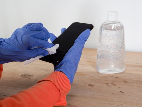 close up of  woman hands wearing blue gloves while cleaning and sanitizing  smartphone  with anti virus bacterial gel against covid-19 , fall dress, orange color over a wood table, earth tones, white background and a gel bottle.