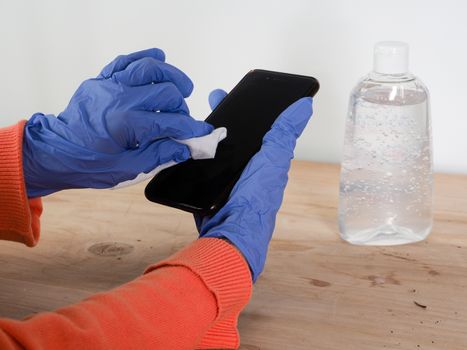 close up of  woman hands wearing blue gloves while cleaning and sanitizing  smartphone  with anti virus bacterial gel against covid-19 , fall dress, orange color over a wood table, earth tones, white background and a gel bottle.