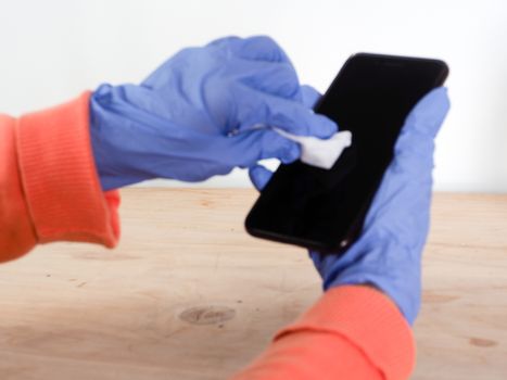 close up of  woman hands wearing blue gloves while cleaning and sanitizing  smartphone  with anti virus bacterial gel against covid-19 , fall dress, orange color over a wood table, earth tones, white background and a gel bottle.