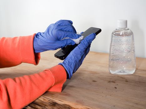 close up of  woman hands wearing blue gloves while cleaning and sanitizing  smartphone  with anti virus bacterial gel against covid-19 , fall dress, orange color over a wood table, earth tones, white background and a gel bottle.