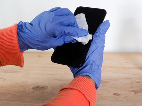 close up of  woman hands wearing blue gloves while cleaning and sanitizing  smartphone  with anti virus bacterial gel against covid-19 , fall dress, orange color over a wood table, earth tones, white background and a gel bottle.