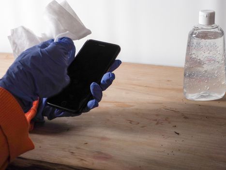 close up of  woman hands wearing blue gloves while cleaning and sanitizing  smartphone  with anti virus bacterial gel against covid-19 , fall dress, orange color over a wood table, earth tones, white background and a gel bottle.