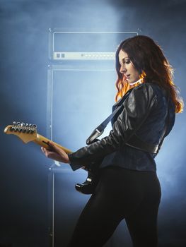 Young beautiful redhead woman playing an electric guitar in front of large amplifier.