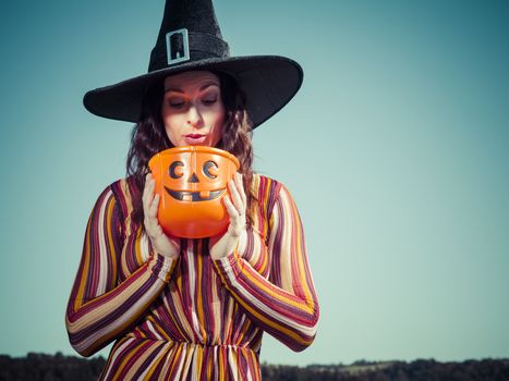 Photo of a young sexy woman holding a pumpkin candy bucket and looking inside and wearing a witch hat for Halloween theme.
