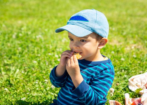 Little boy eating a biscuit on park lawn, toddler wearing blue cap.
