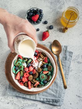 Trendy food - pancake cereal. Milk pouring on heap of multicolor mini cereal pancakes in bowl. Tiny cereal pancakes with berries in craft plate over gray cement background. Vertical. Top view,flat lay