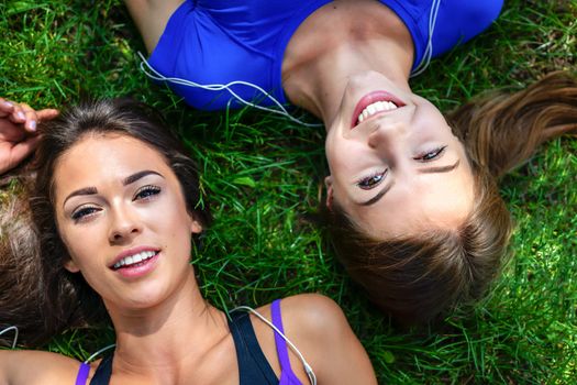 Top view of two beautiful and young girls rest on the grass after outdoor workout in a sunny day