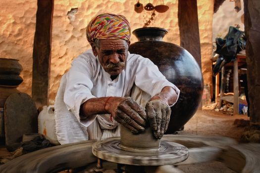 Indian potter at work: throwing the potter's wheel and shaping ceramic vessel and clay ware: pot, jar in pottery workshop. Experienced master. Handwork craft from Shilpagram, Udaipur, Rajasthan, India