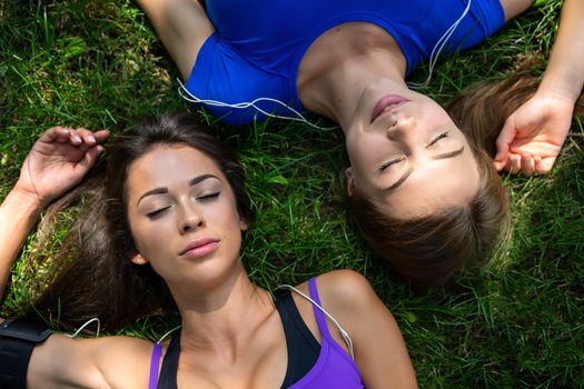 Top view of two beautiful and young girls resting with close eyes on the grass after outdoor workout in a sunny day