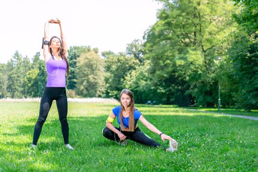 Healthy lifestyle concept - two young and fit girls stretching before jogging on the grass in a park on a sunny morning.
