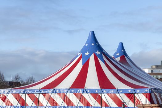 Red and white circus tent topped with bleu starred cover against a sunny blue sky with clouds