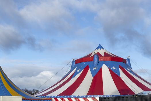 Red and white circus tent topped with bleu starred cover against a sunny blue sky with clouds