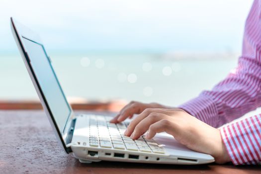 Woman hand using laptop to work study on work desk with clean nature beach outdoor background. Business, financial, trade stock maket and social network concept.