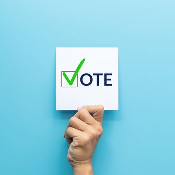 hand holding white paper with the "vote" and green check mark voting symbols in checkbox of the inscription isolated on blue background