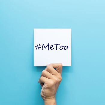 hashtag #MeToo on white paper in hand isolated on blue background. #MeToo is a campaign for movement against sexual harassment and sexual assault.