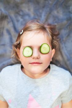 Funny small girl with piece of cucumber on their eyes like a mask, beauty and health concept, indoor closeup portrait
