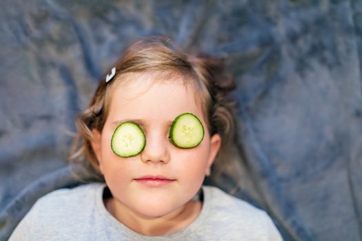 Funny small girl with piece of cucumber on their eyes like a mask, beauty and health concept, indoor closeup portrait