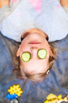 Funny small girl with piece of cucumber on their eyes like a mask, beauty and health concept, indoor closeup portrait